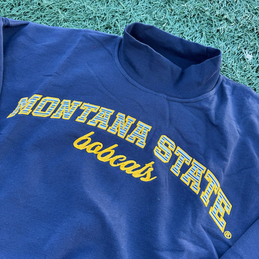 A close-up of a royal blue mockneck crewneck with 'MONTANA STATE' and 'bobcats' printed in yellow across the chest, laid flat on green artificial turf.