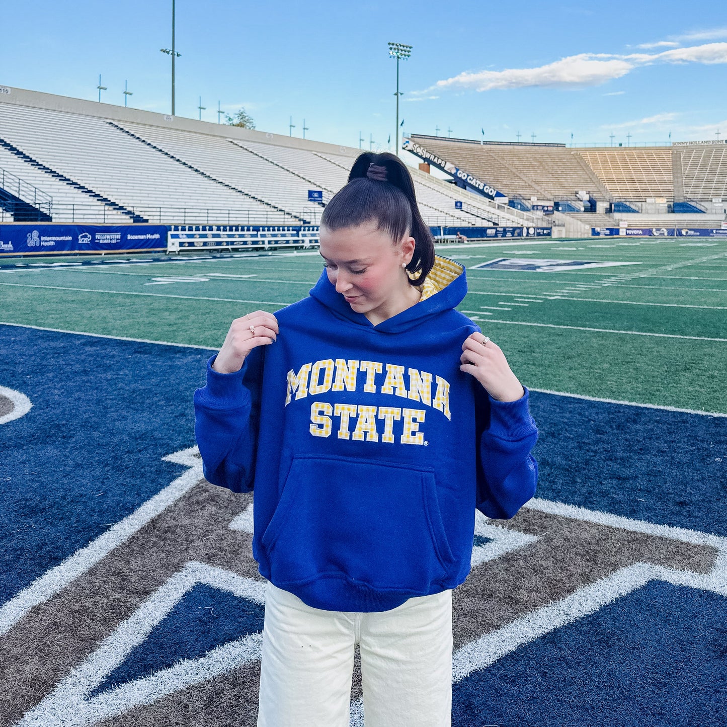 A woman stands on a football field wearing a royal blue Montana State hoodie and white pants, with stadium seating and blue sky in the background.