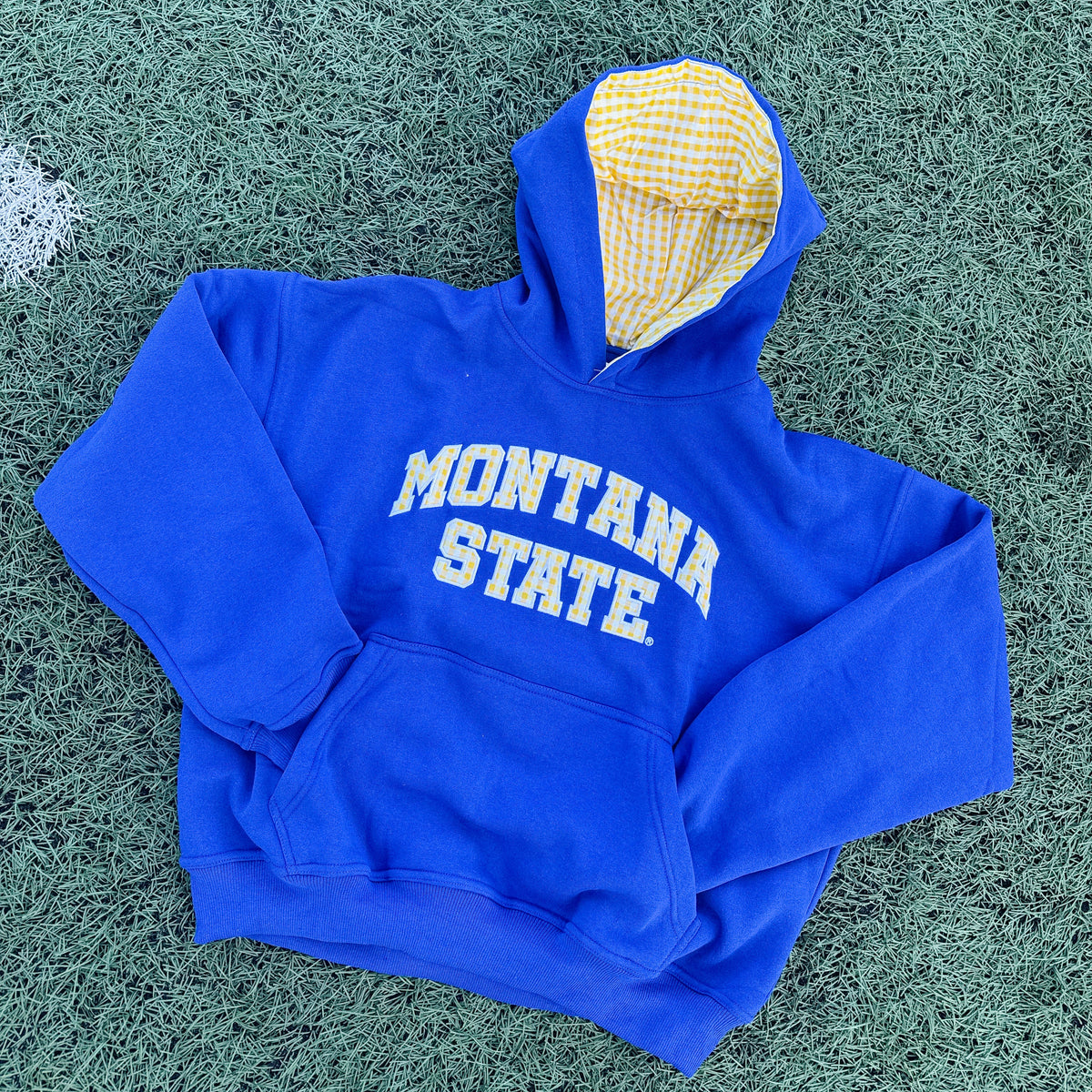 A royal blue Montana State hoodie with yellow gingham varsity lettering, laid flat on green turf. The hood is open, revealing a yellow and white gingham lining.