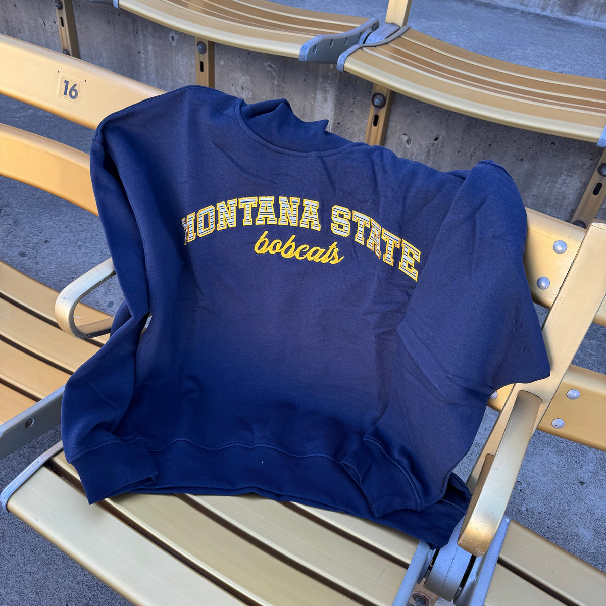 A navy blue mockneck sweatshirt with 'MONTANA STATE bobcats' printed in yellow across the chest, resting on a gold stadium seat.