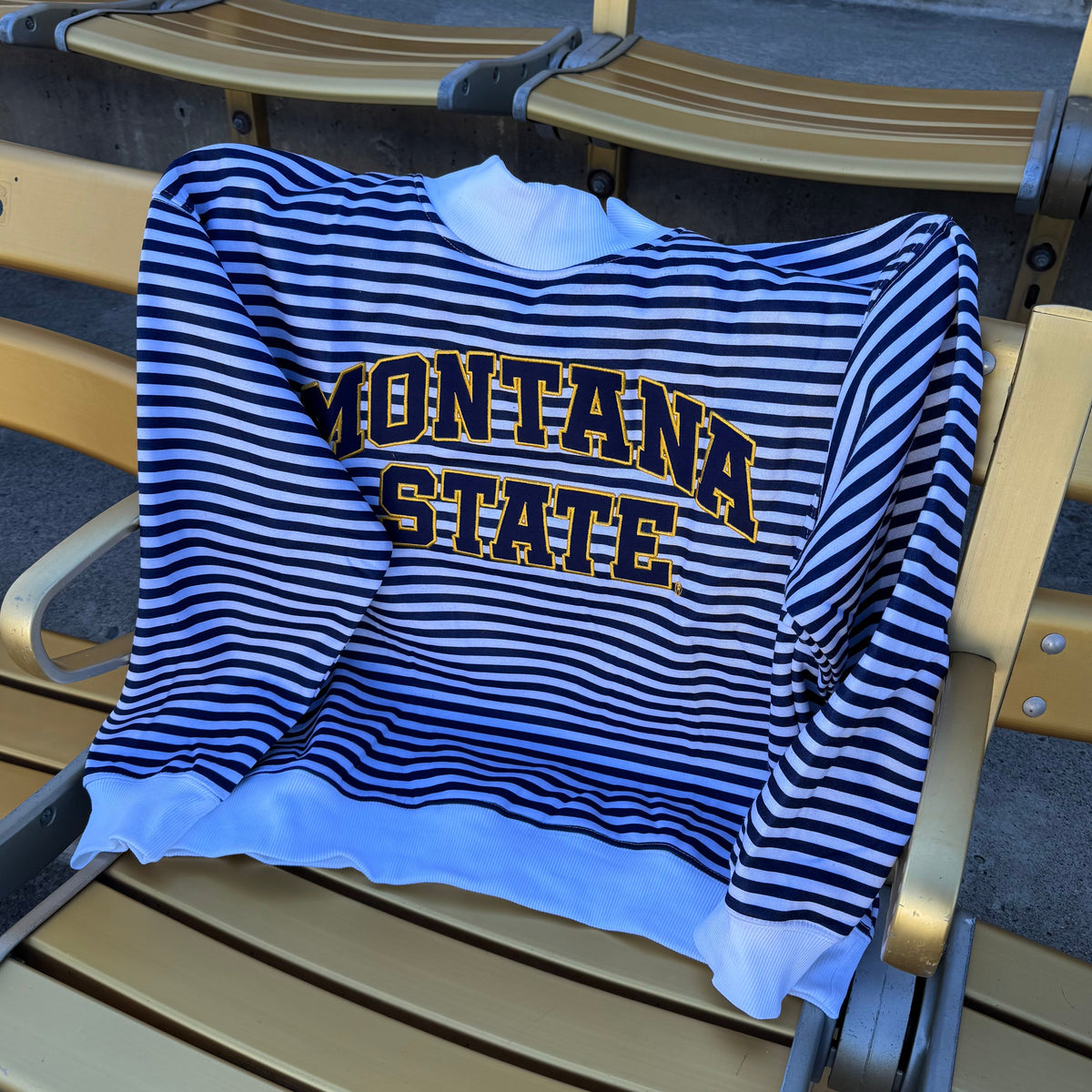 A blue and navy striped long-sleeve sweatshirt with 'MONTANA STATE' printed in gold across the chest, draped over a gold stadium seat.