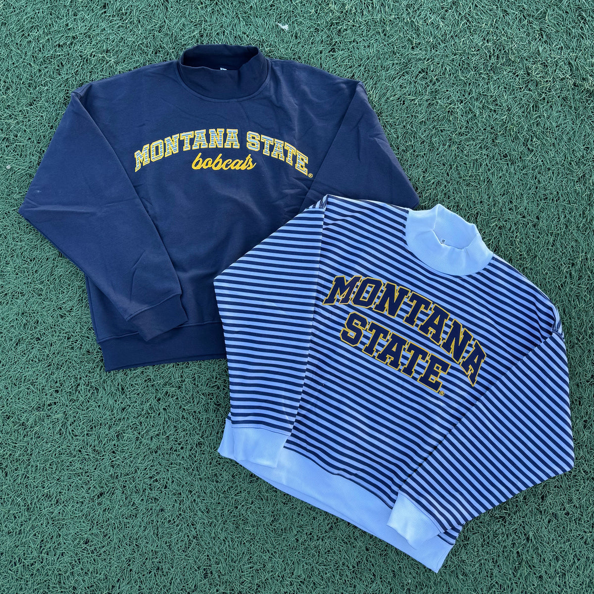 Two Montana State Bobcats sweatshirts laid flat on green artificial turf. One is navy blue with yellow text, and the other is navy and white striped with navy and yellow text.