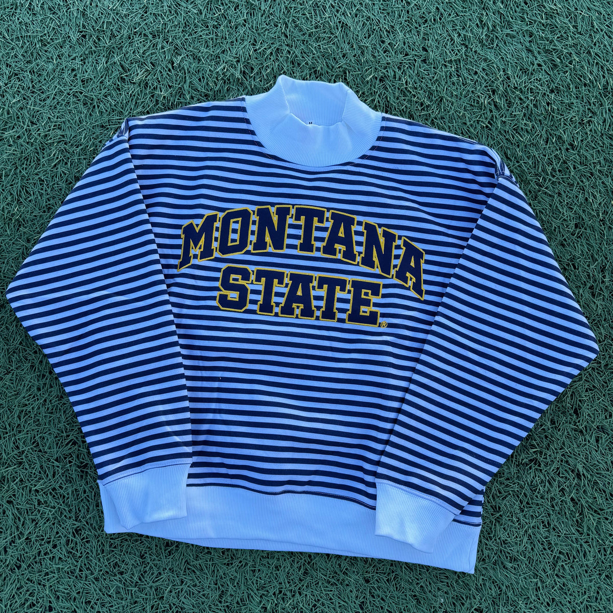A blue and white horizontally striped mockneck sweatshirt with 'MONTANA STATE' printed across the chest in navy and gold, laid flat on green grass.