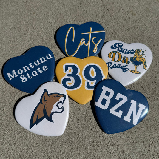 Five heart-shaped pin-back buttons arranged on ground showing off diffrent designs.