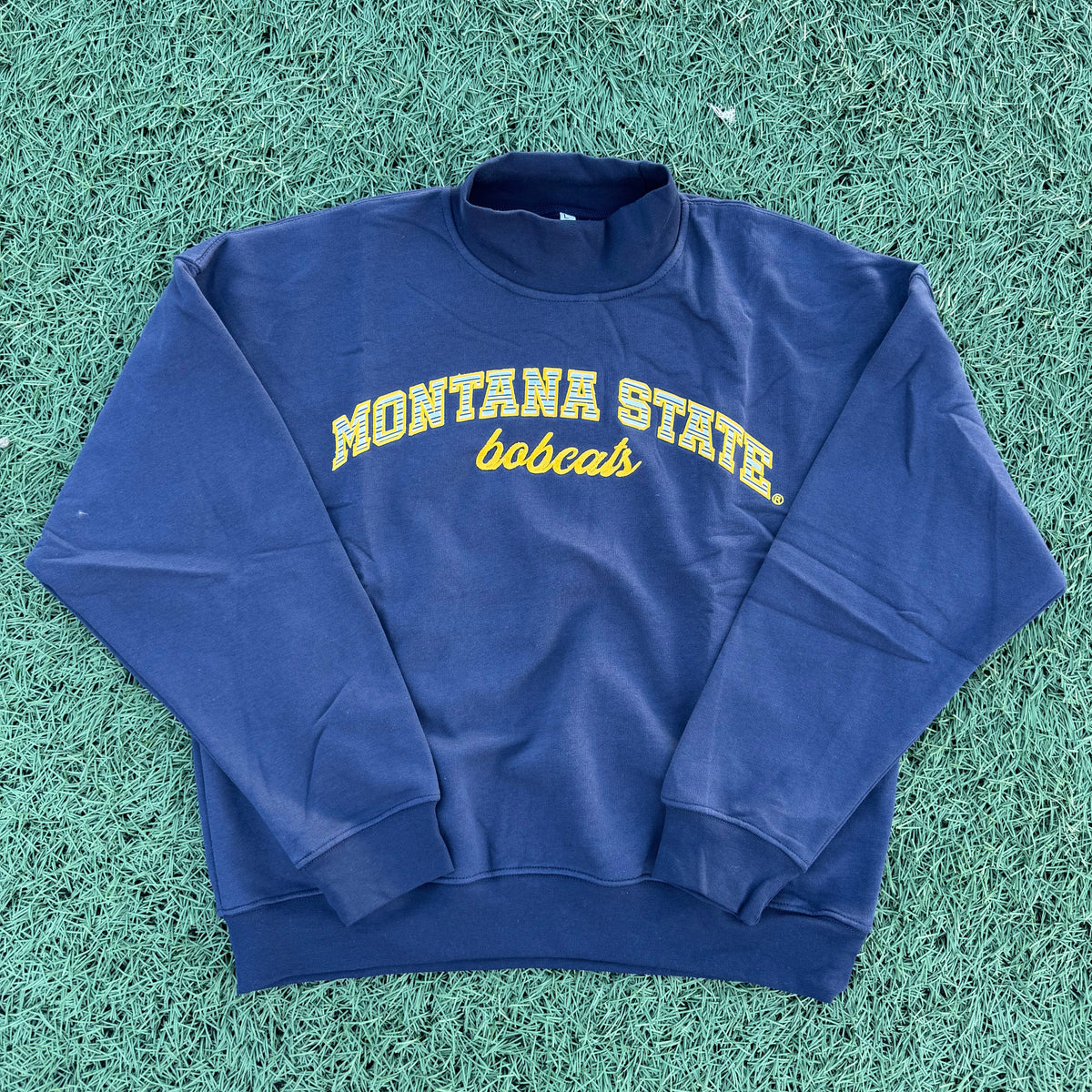 A navy blue long-sleeve sweatshirt with 'MONTANA STATE Bobcats' embroidered in yellow stripes on the front, laid flat on green artificial turf.