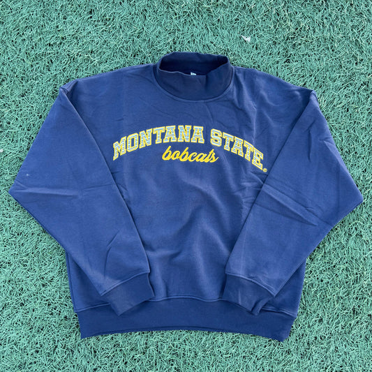 A navy blue long-sleeve sweatshirt with 'MONTANA STATE Bobcats' embroidered in yellow stripes on the front, laid flat on green artificial turf.